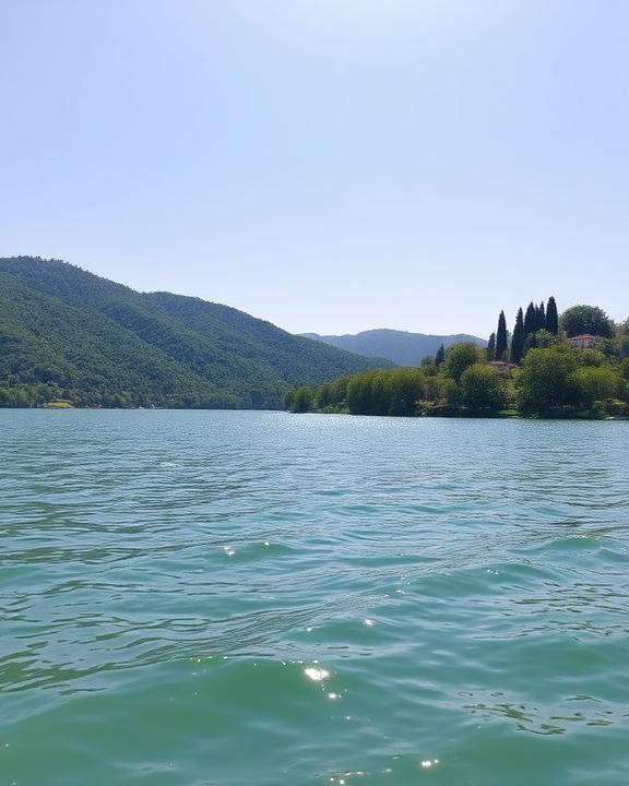 A serene and picturesque view of Lake Trasimeno in Italy during a bright sunny day, with gentle waves lapping against the shore, surrounded by lush green hills and trees.